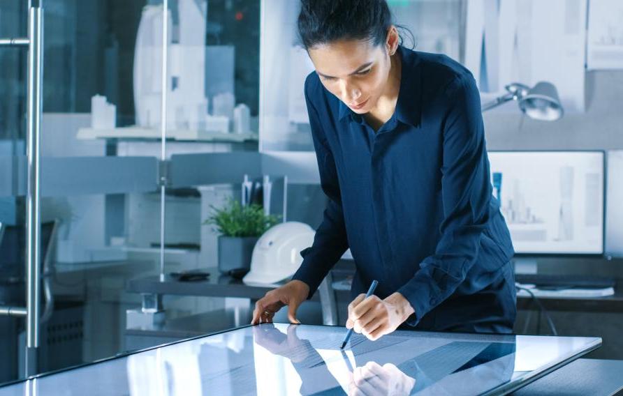 Talented Young Female Architectural Designer Draws Building Concept on a Graphics Tablet Display. Clean Minimalistic Office, Concrete Walls Covered by Blueprints.